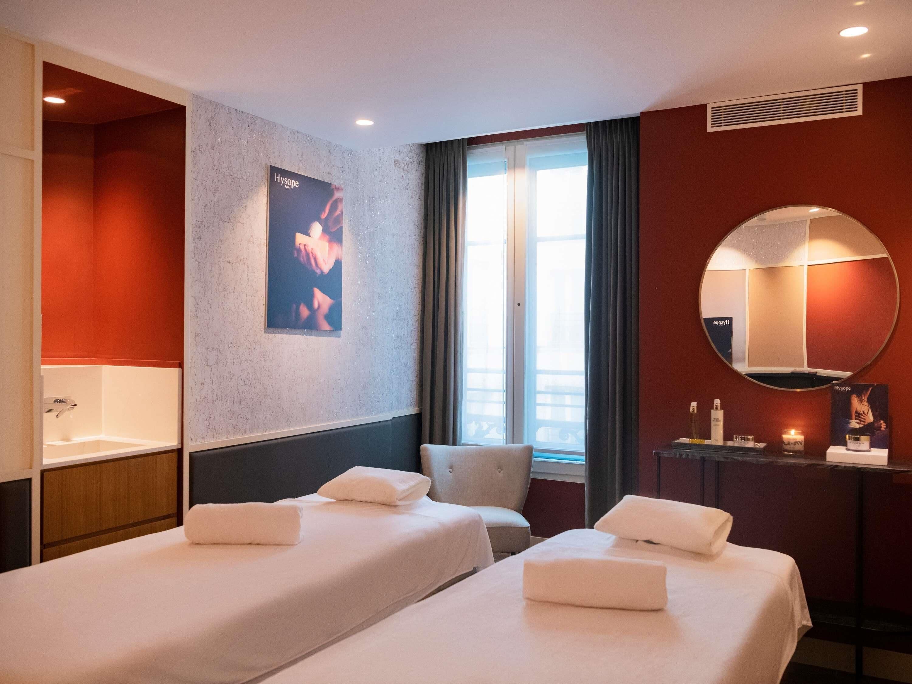 Une chambre d’hôtel avec deux lits, un grand miroir, une affiche au mur et une fenêtre avec des rideaux.