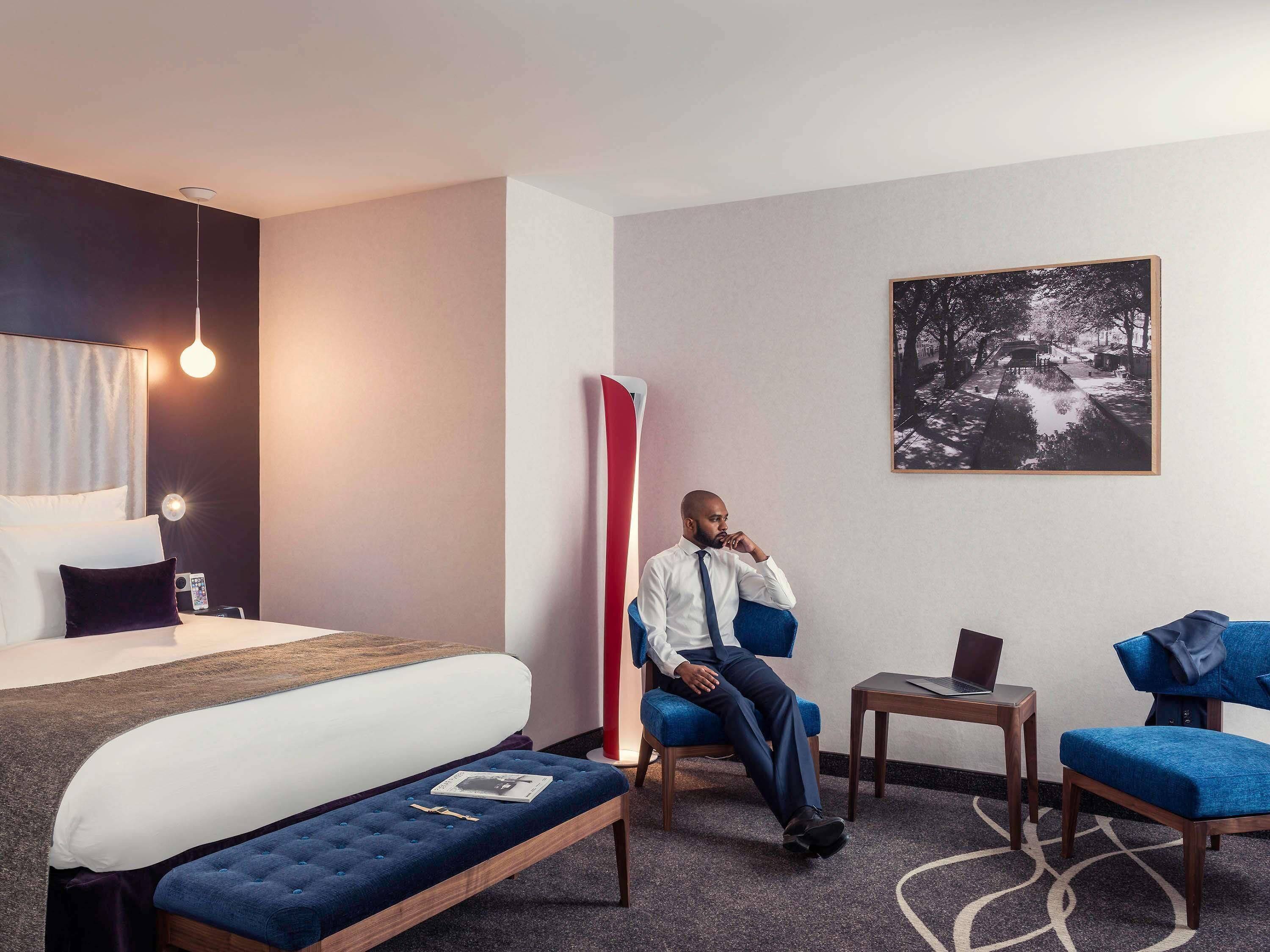 Une chambre d’hôtel comprenant un lit, un coin salon avec un fauteuil et une table, un bureau avec un ordinateur portable et un tableau accroché au mur.