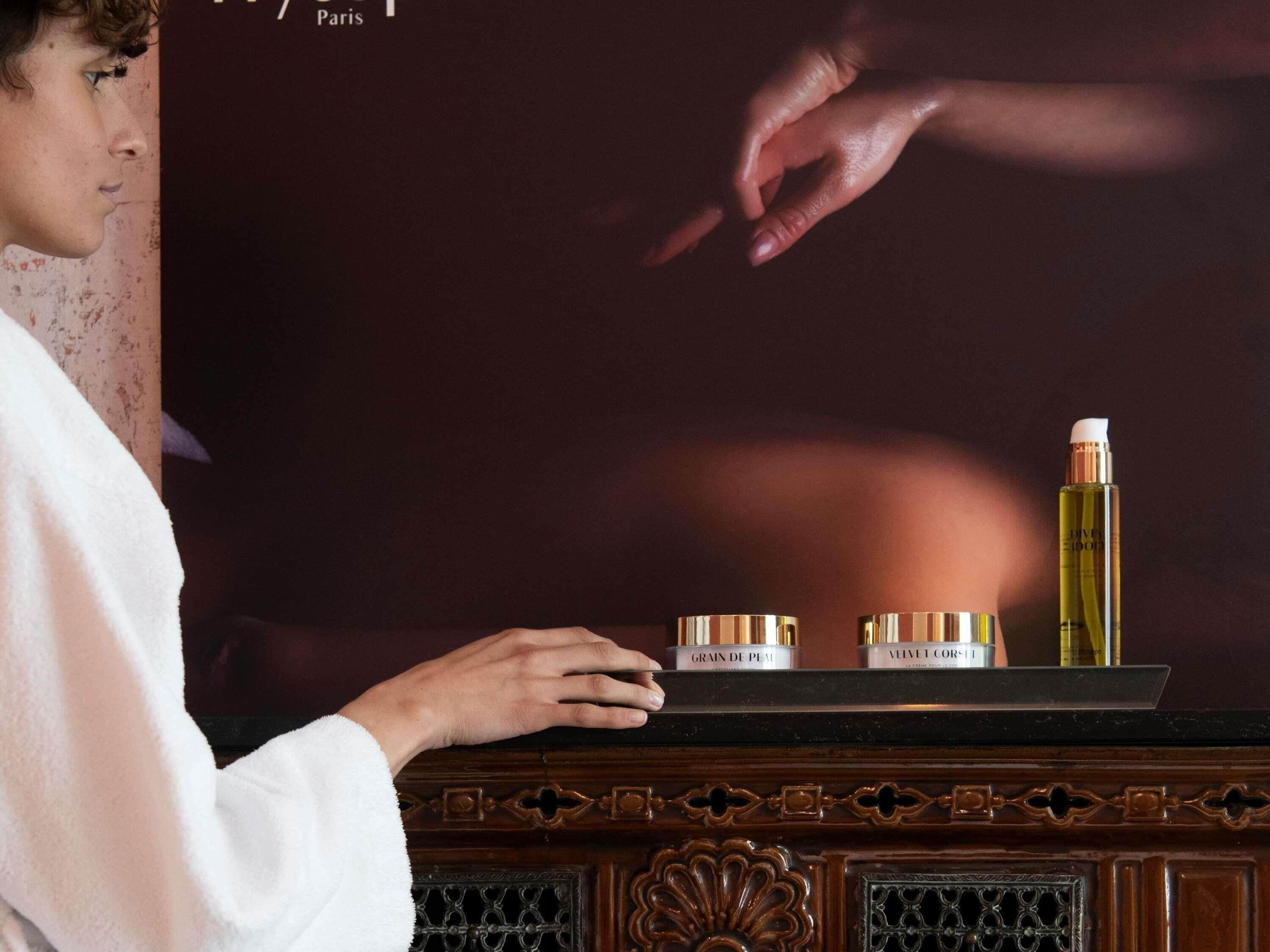 A person in a white robe standing in front of a wooden cabinet with three cosmetic containers labeled "Grain de Peau," "Velvet Corset," and "Divy Elixir."
