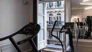A home gym with a treadmill and elliptical, a balcony with a view of a building, and a large mirror reflecting the gym equipment. - Mercure Paris Opéra Garnier (Paris)