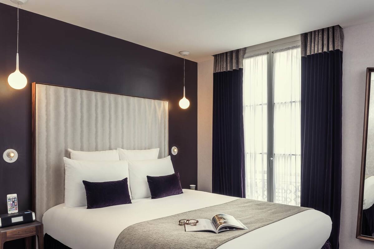 A hotel room with a large bed, two bedside tables, a bedside lamp, a window with curtains, and a mirror on the wall.