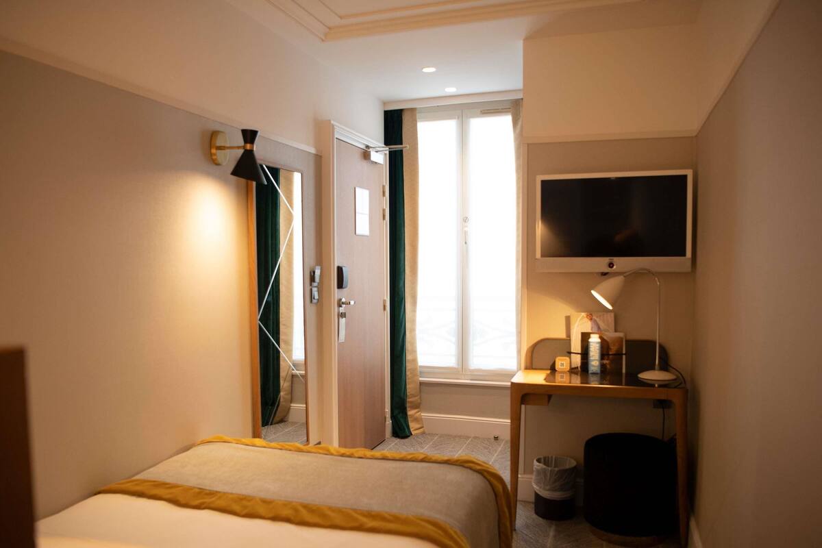 A hotel room with a bed, a television, a desk with a lamp, a trash bin, and a window with curtains.