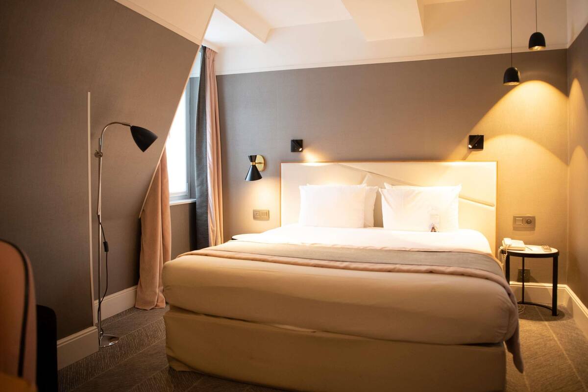 A hotel room with a large bed, a bedside table, a lamp, and a window with curtains.