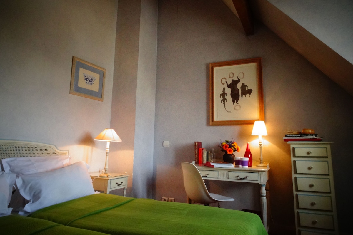 A bedroom with a bed, a desk, a chair, and a dresser. There are two lamps on the desk and a framed picture on the wall.