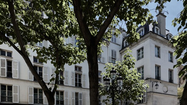 Une rue bordée d’arbres, avec un immeuble aux façades blanches et aux volets colorés.