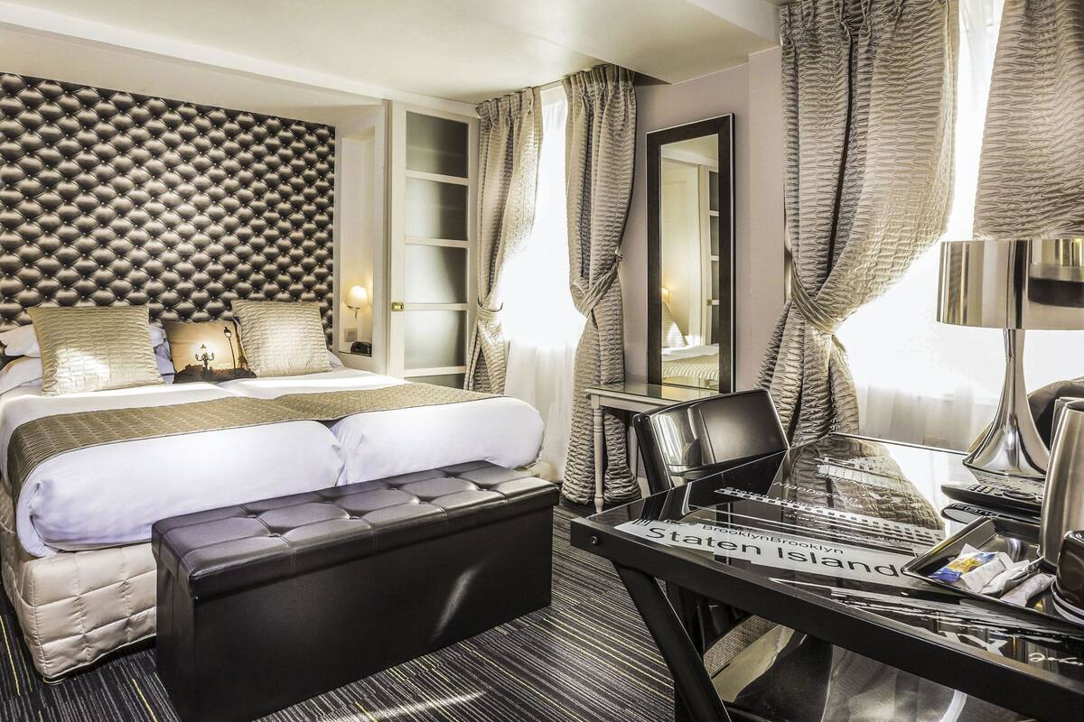 A hotel room with a tufted headboard, a bed with white linens, a black leather ottoman, a desk with a laptop, and a large mirror.