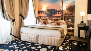 A hotel room with a bed, a desk, a chair, a lamp, and a view of a cityscape. - Hotel Ile de France Opéra (Paris)