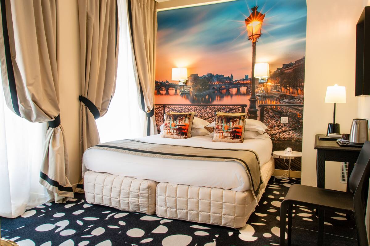 A hotel room with a bed, a desk, a chair, a lamp, and a view of a cityscape.