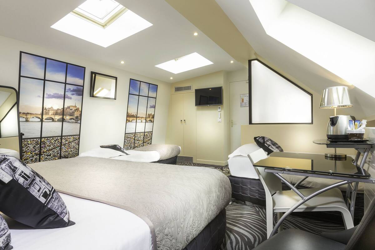 A modern hotel room with a large bed, a desk with a lamp, a TV, and a view of a cityscape through a window.