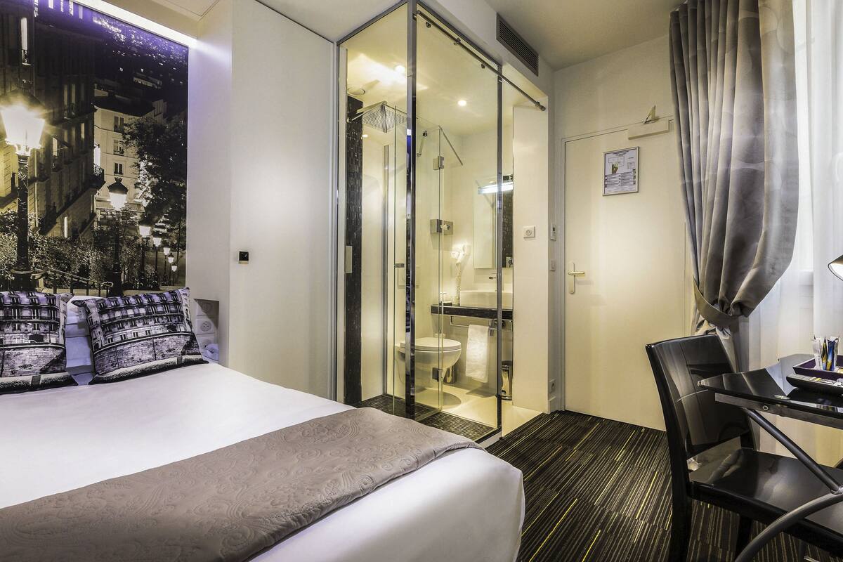A hotel room with a bed, a desk, a chair, a shower, and a view of a cityscape through a glass door.