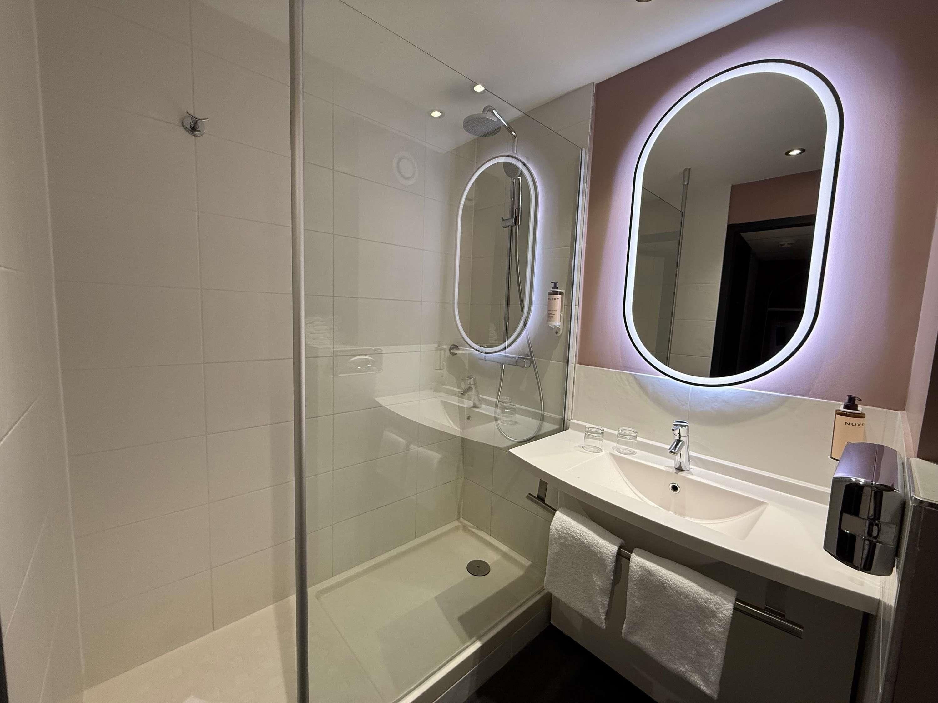 A modern bathroom with a glass-enclosed shower, a white sink with a mirror, and a Nuxe bottle on the counter.