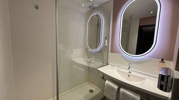 A modern bathroom with a glass-enclosed shower, a white sink with a mirror, and a Nuxe bottle on the counter.
