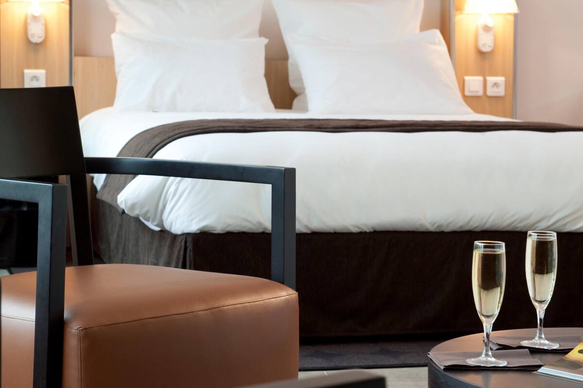 Ein Hotelzimmer mit einem Bett, zwei Nachttischlampen, einem Sessel und zwei Sektgläsern auf einem kleinen Tisch.