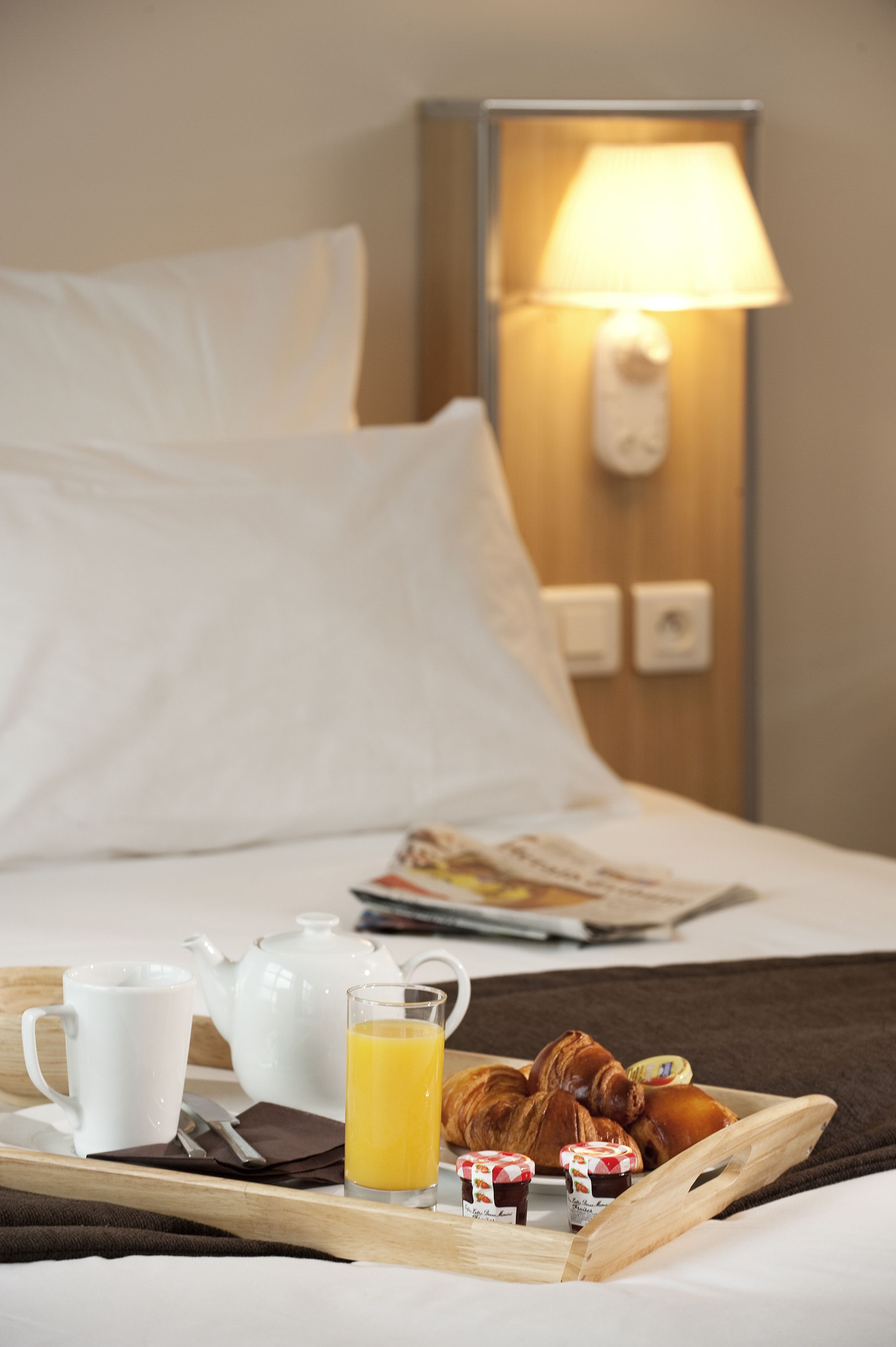 A neatly made bed with white linens, a bedside lamp, a tray with breakfast items including croissants, a teapot, a cup, and a glass of orange juice.