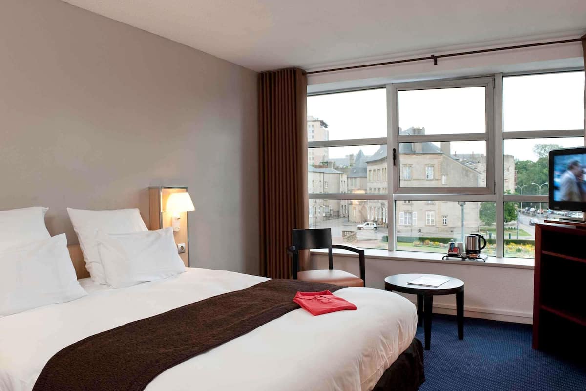 A hotel room with a large bed, a TV, a small table, and a window with a view of buildings.