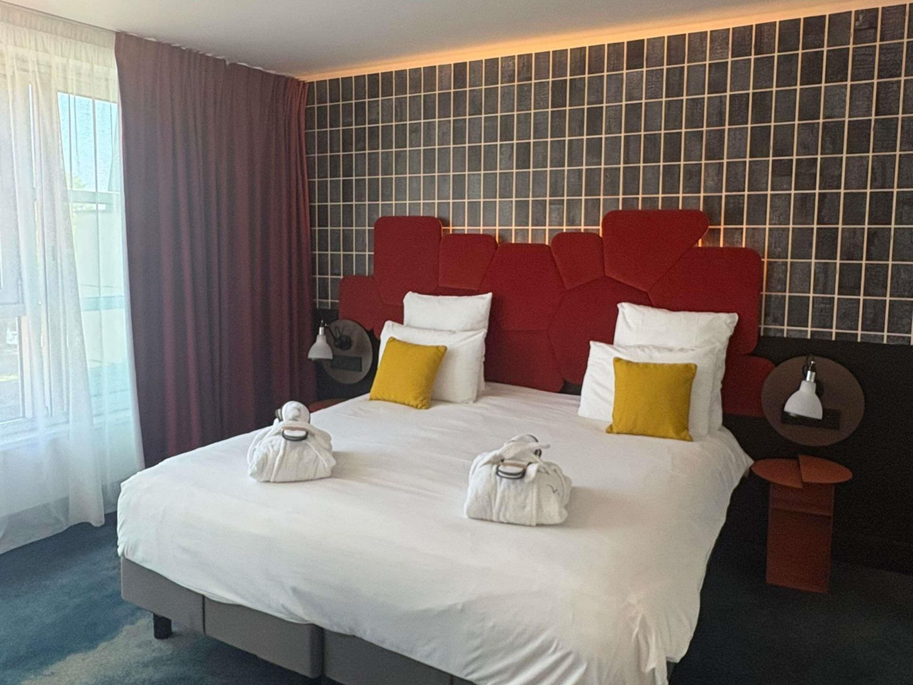 Habitación de hotel con una cama grande, cabecera roja y vistas al exterior a través de una ventana con cortinas.