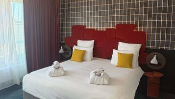 Habitación de hotel con una cama grande, cabecera roja y vistas al exterior a través de una ventana con cortinas.