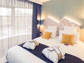 Quarto de hotel com uma cama grande, duas almofadas, cabeceira estampada com flores e janela com cortinas leves.