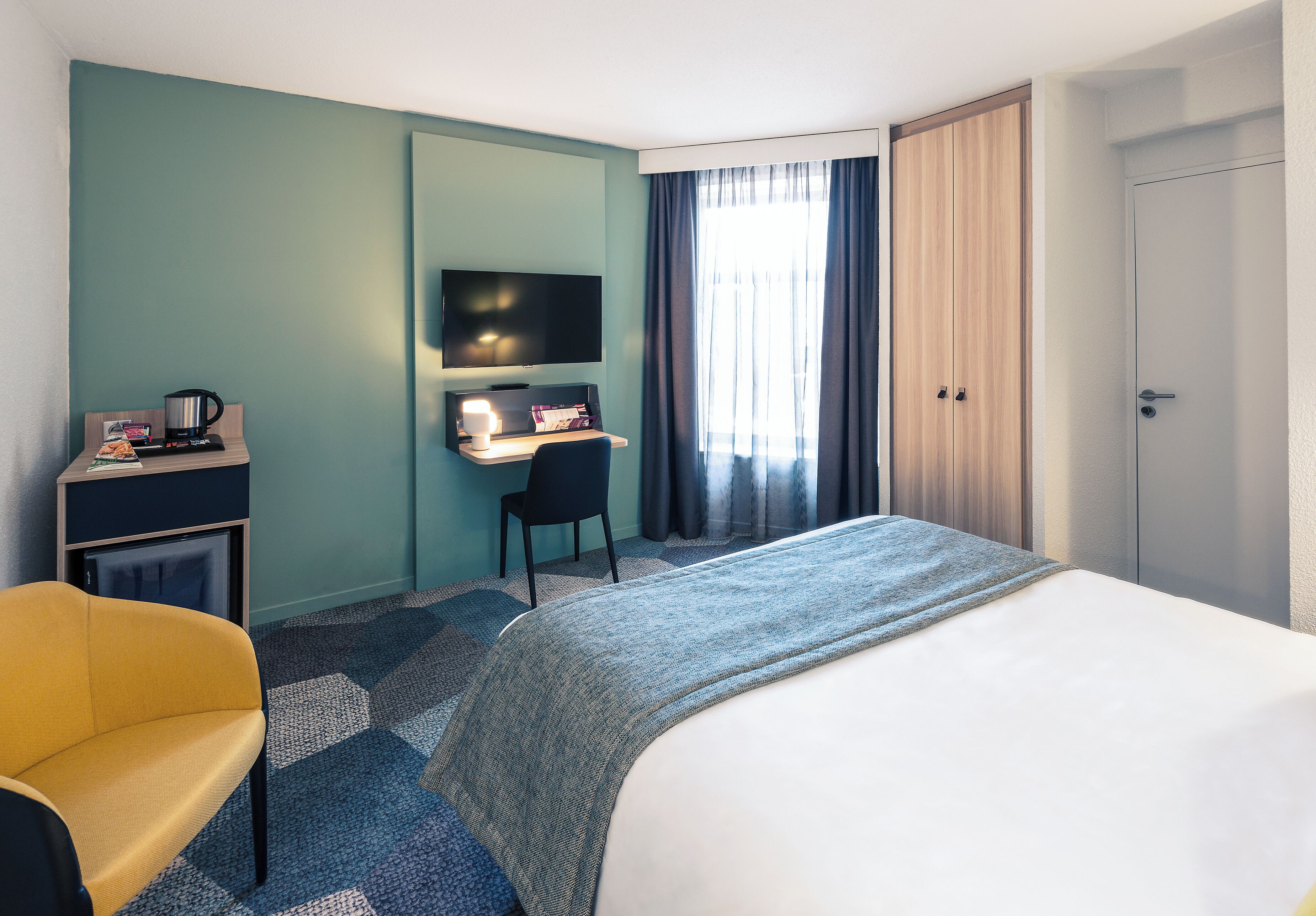 A hotel room with a bed, a desk with a computer, a chair, a television, and a wardrobe.