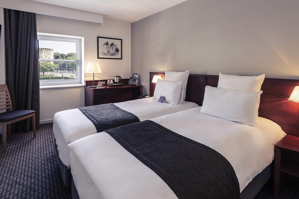 A hotel room with two beds, a desk, a chair, a window with a view of greenery, and a framed picture on the wall.