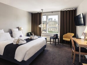 Habitación de hotel con cama, escritorio, silla, televisor y ventana con cortinas.