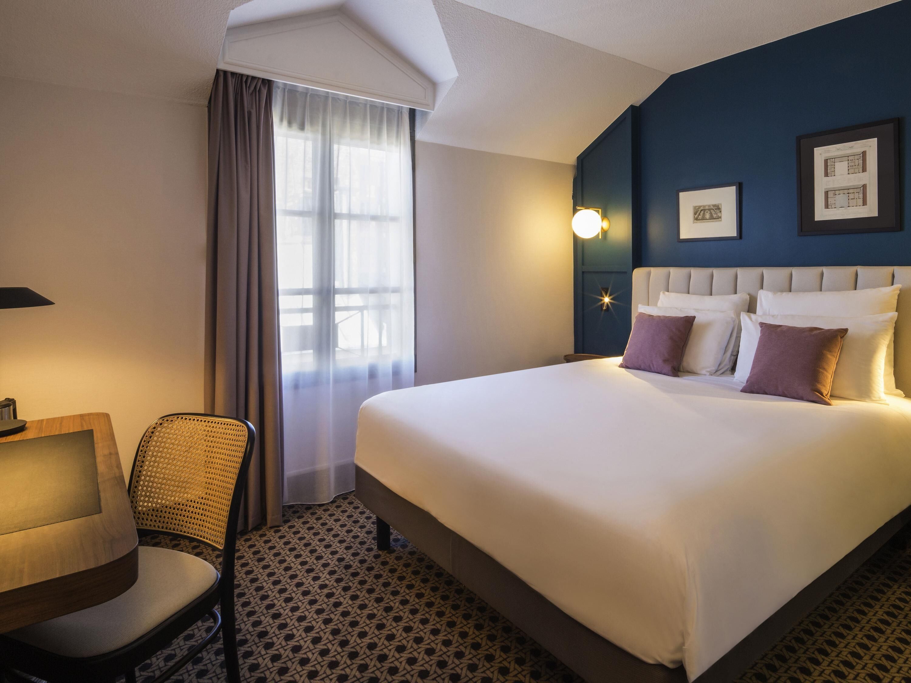 A hotel room with a large bed, a chair, a desk, and a window with curtains.