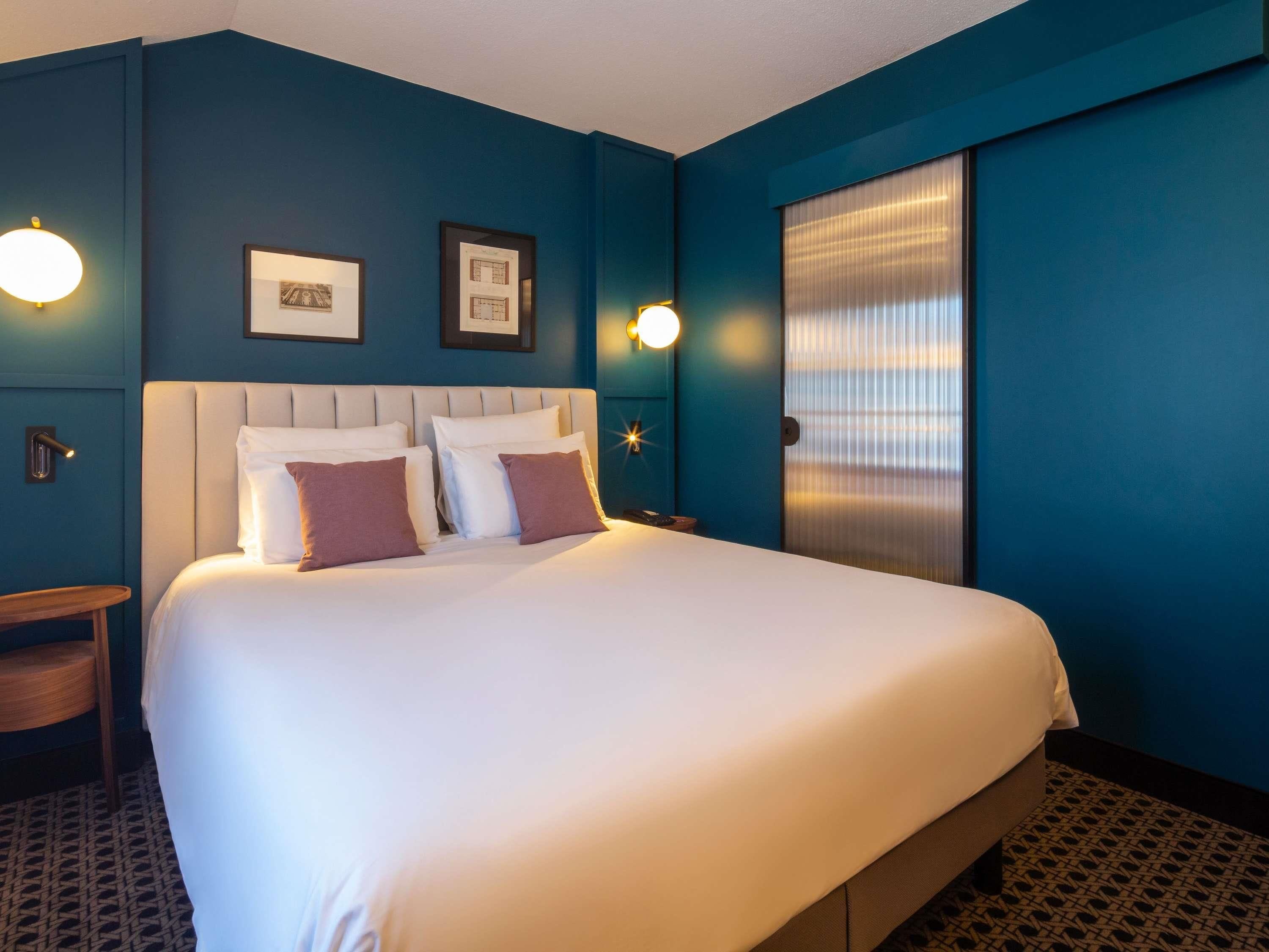 Ein Hotelzimmer mit einem großen Bett, zwei gerahmten Bildern an der Wand, einem Nachttisch und einem Fenster mit Jalousien.