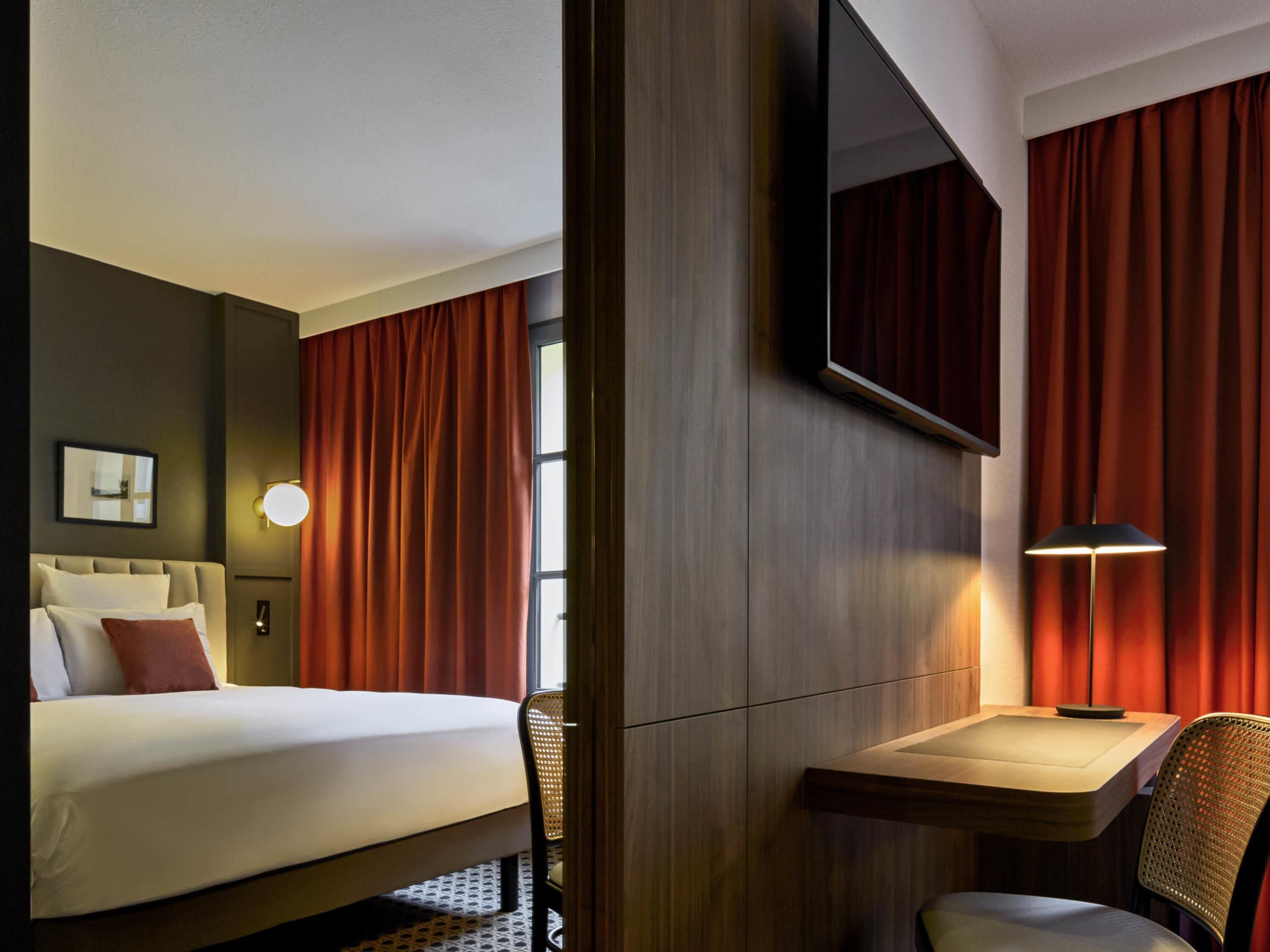 A hotel room with a bed, a desk, a chair, a mirror, and a window with curtains.