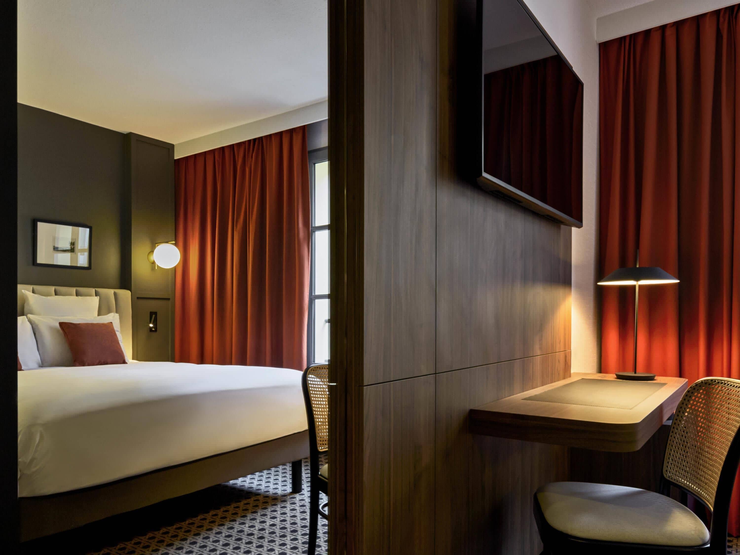 A hotel room with a bed, a desk, a chair, a lamp, and a mirror.