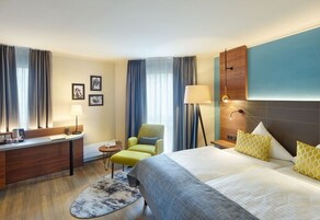 Habitación de hotel con cama, escritorio, silla, lámpara y una pequeña mesa.
