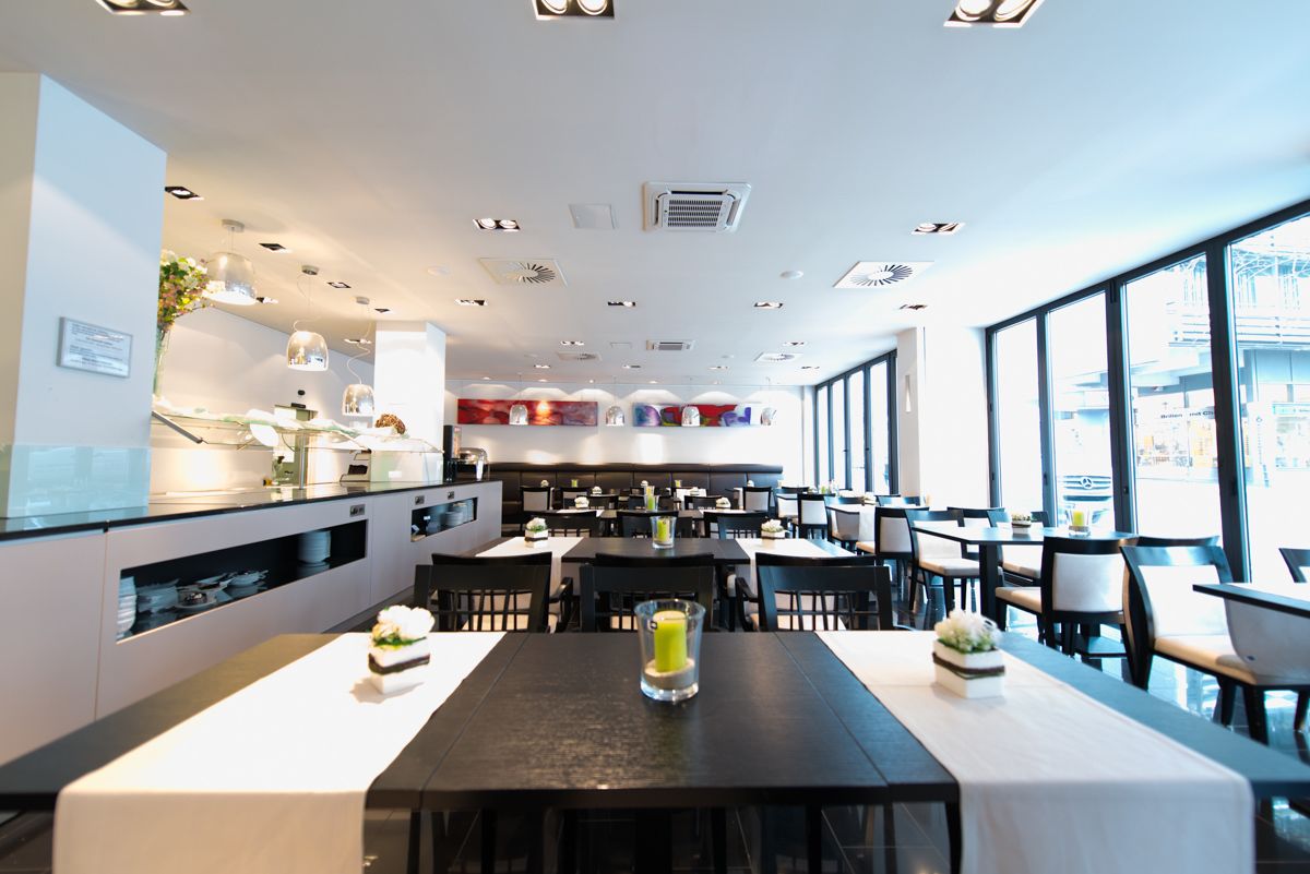 A modern restaurant with a long dining table, multiple seating areas, and large windows offering a view of the city.