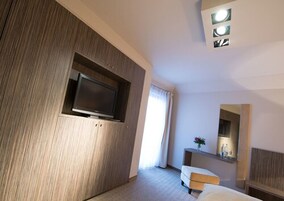 Una habitación de hotel moderna con un televisor de pantalla plana montado en un panel de madera, una mesita de noche con un florero y un espejo grande encima.