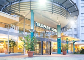 The exterior of the Tierpark Hotel with a glass facade, a curved entrance, and potted plants in front.