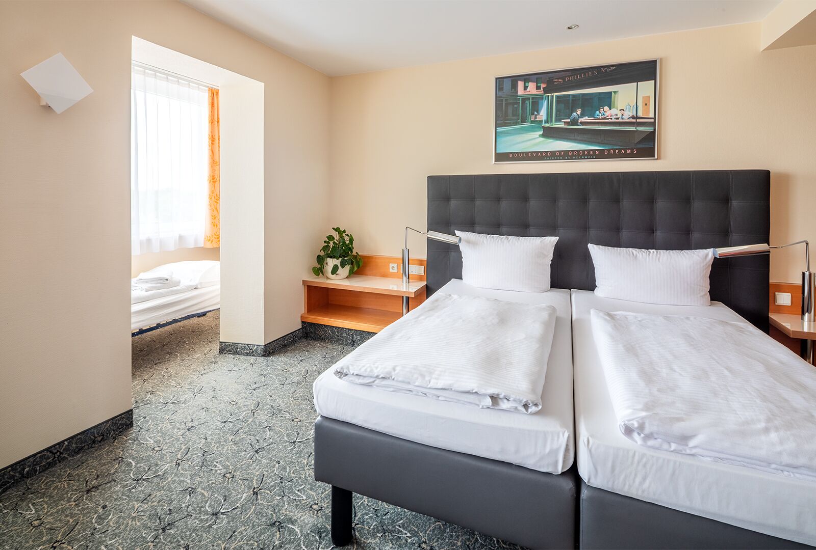 A hotel room with a large bed, a bedside table, a potted plant, a window with curtains, and a painting on the wall.