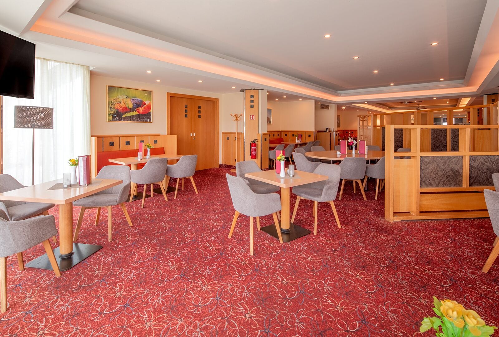 A hotel room with a red patterned carpet, multiple seating areas with tables and chairs, a television, and a painting on the wall.