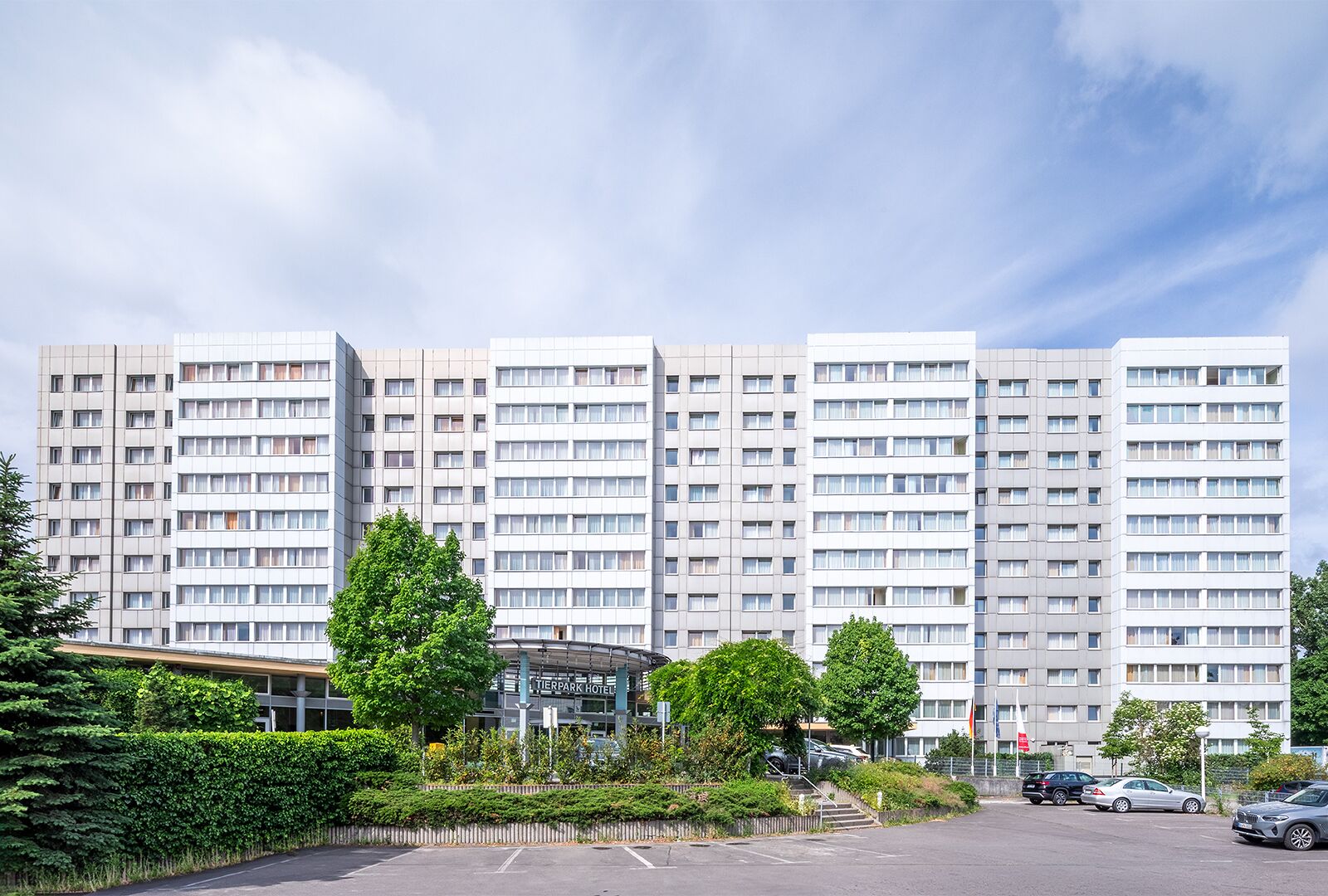 A multi-story hotel with a modern design, featuring a central entrance and a parking area.