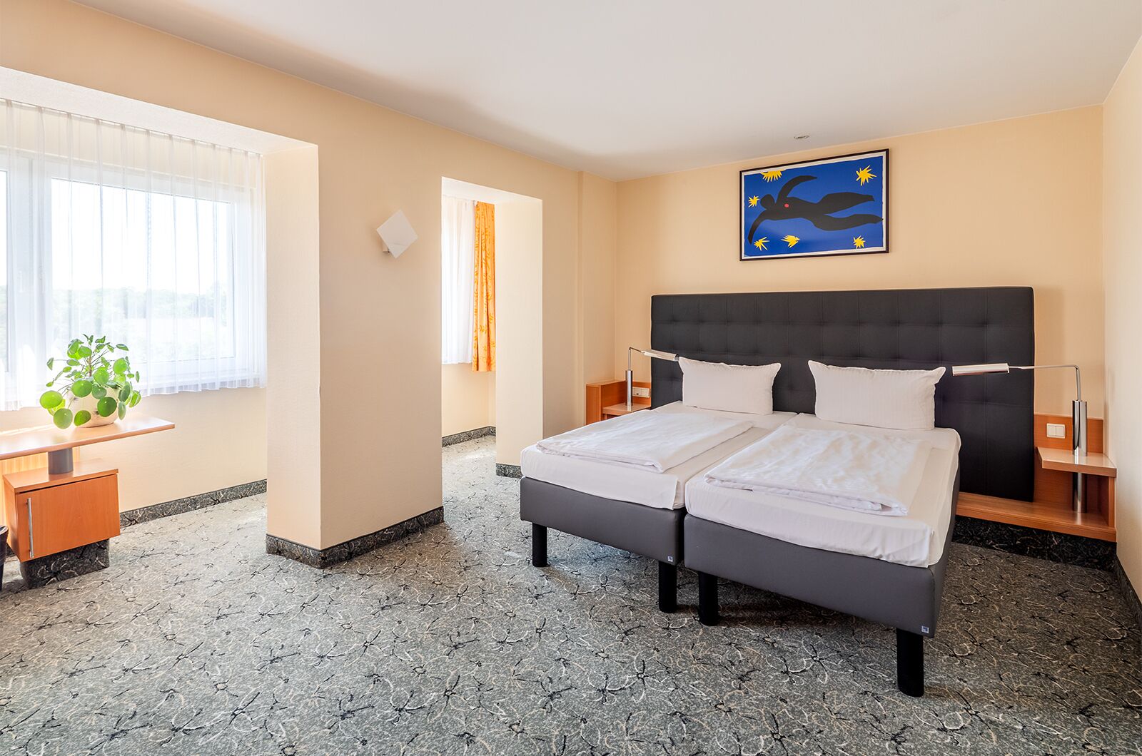 A hotel room with a bed, bedside tables, a desk, a chair, a potted plant, and a wall with a flag.