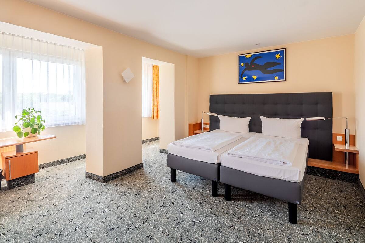 A hotel room with a bed, bedside tables, a desk, a chair, a potted plant, and a wall with a flag.