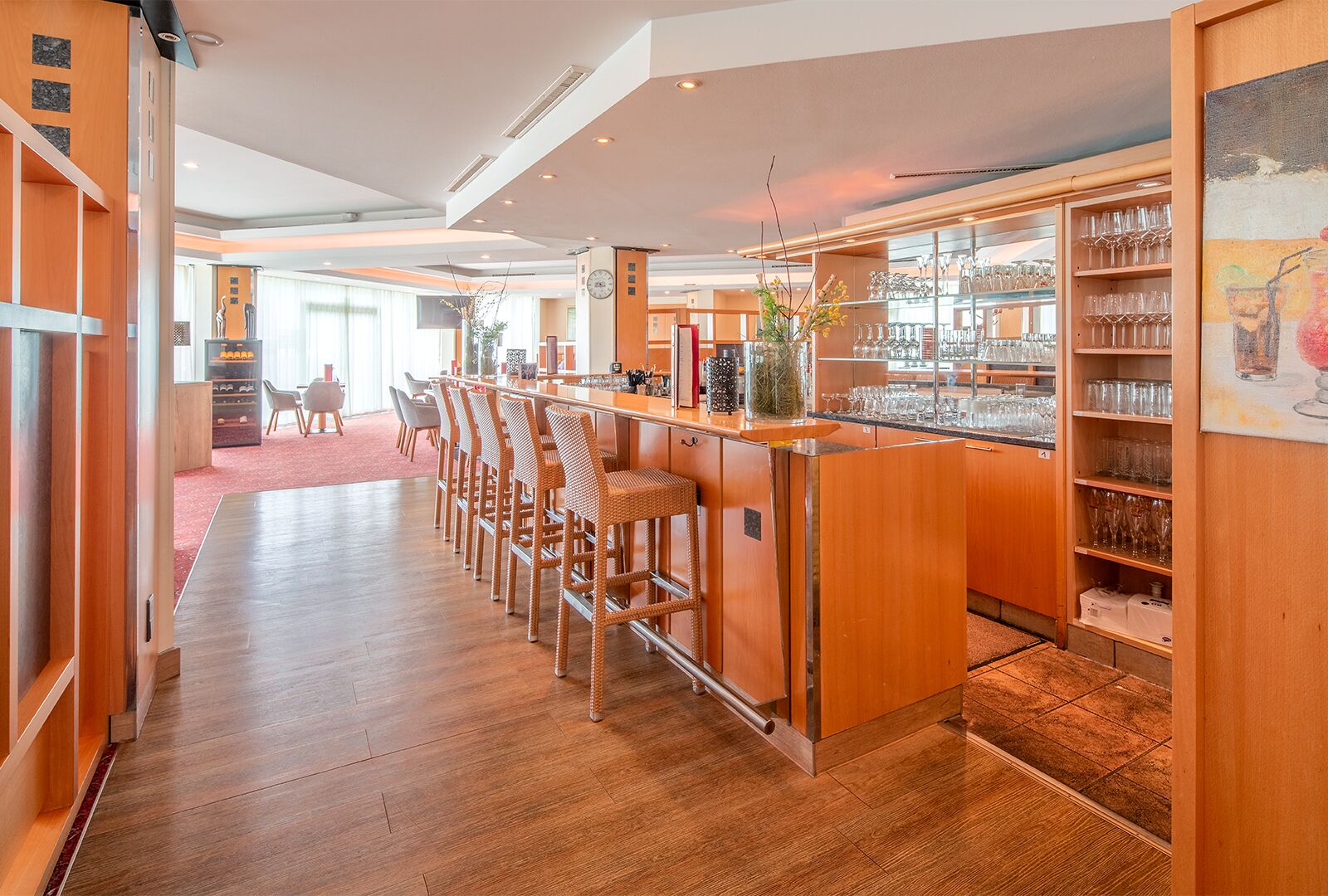 Una zona de bar con taburetes de madera y una barra, un área de asientos con sillas, y una vitrina de vidrio con varios vasos.