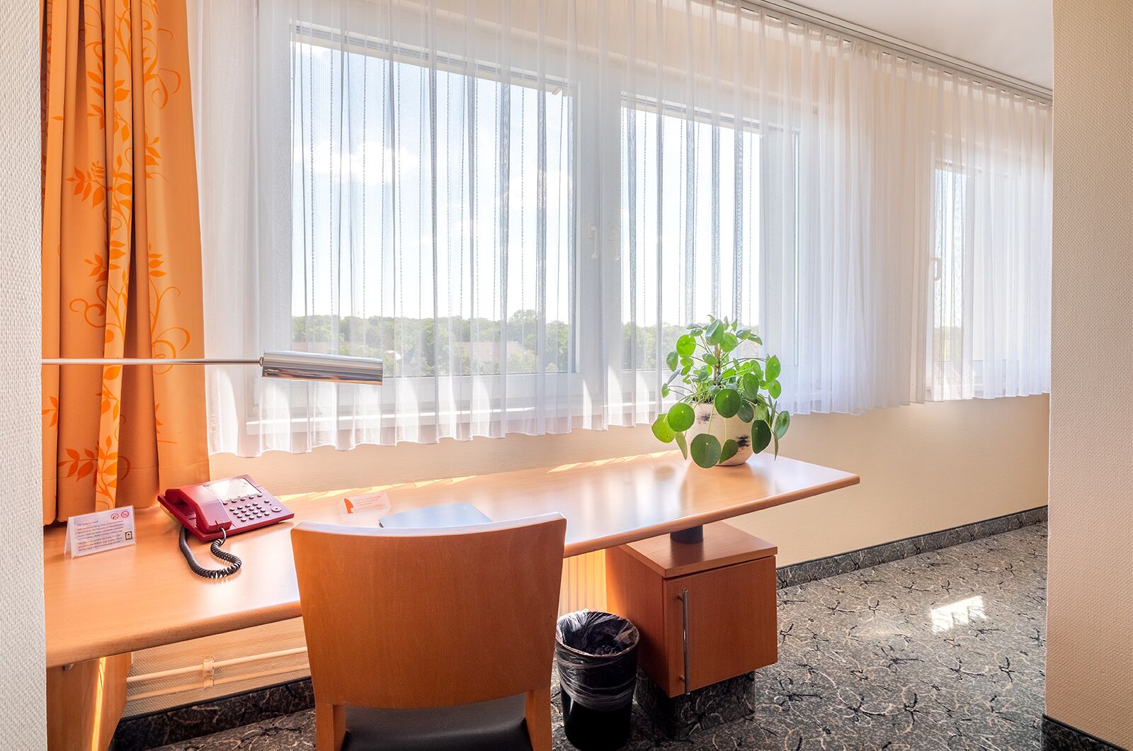 Habitación de hotel con un escritorio de madera, teléfono, cesto de basura, planta en maceta y ventana con cortinas traslúcidas.