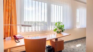 Habitación de hotel con un escritorio de madera, teléfono, cesto de basura, planta en maceta y ventana con cortinas traslúcidas.