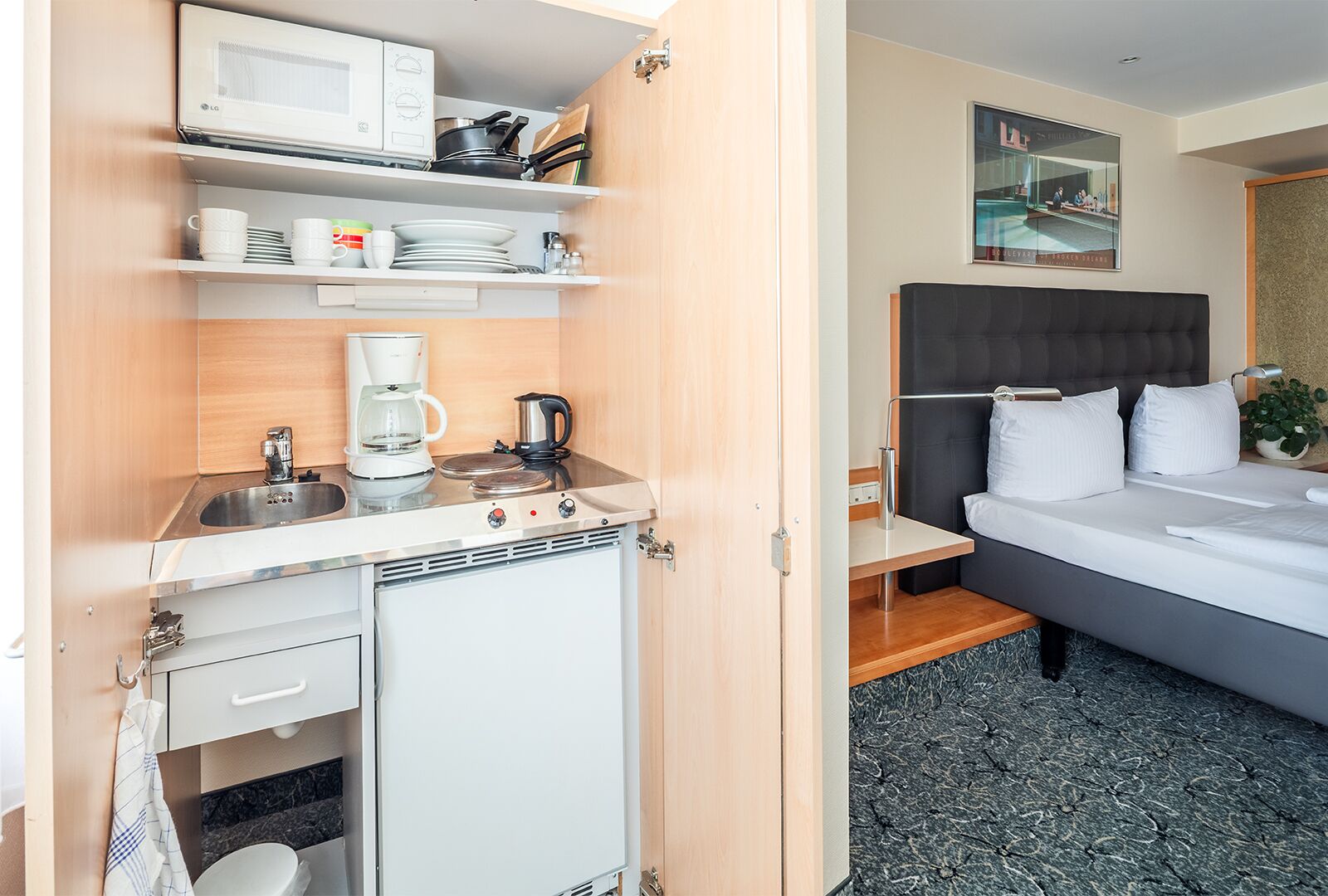 Una habitación de hotel compacta con kitchenette, una cama con ropa de cama blanca, una mesita de noche pequeña y un cuadro en la pared.