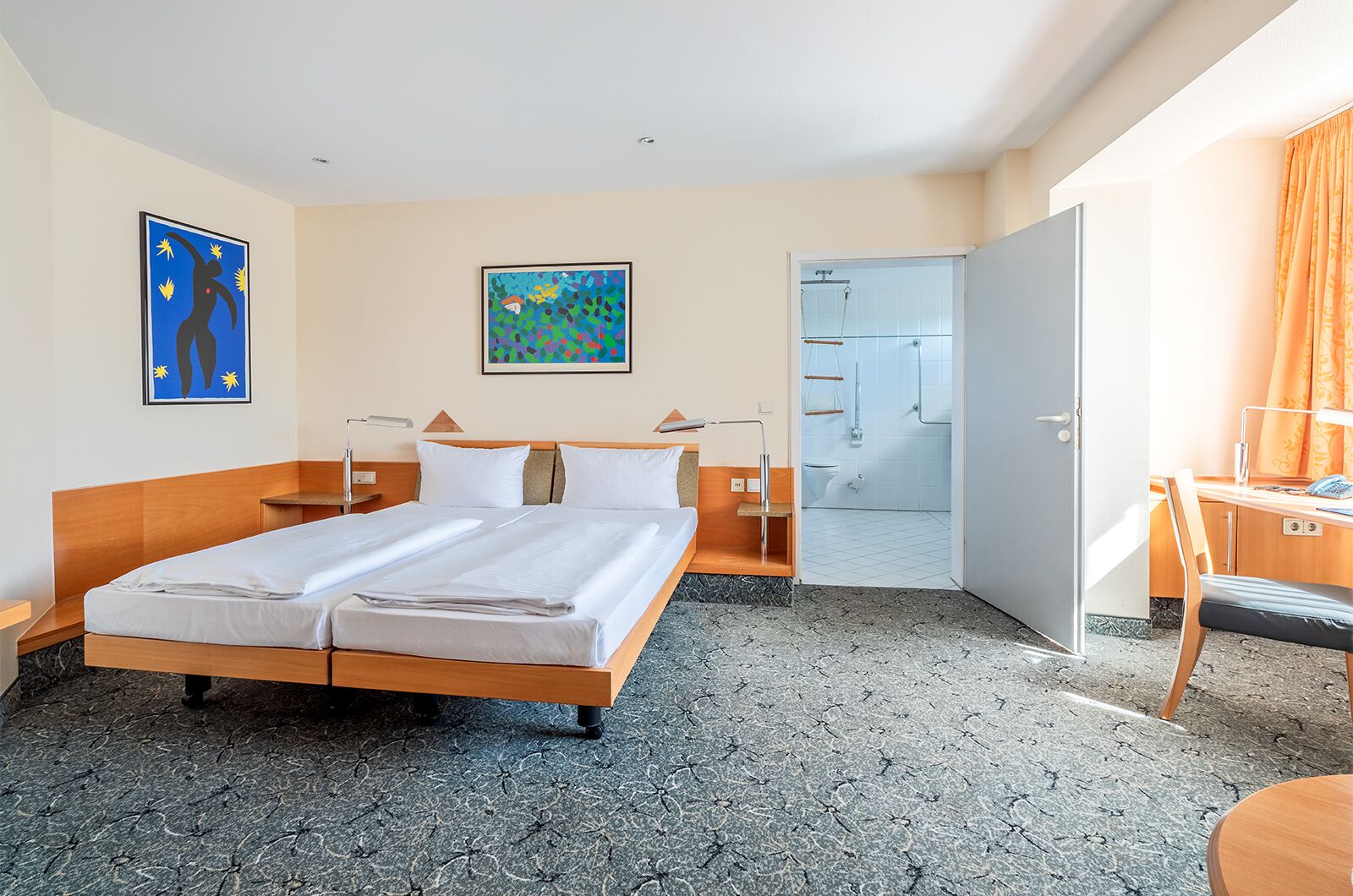 Ein Hotelzimmer mit Bett, Nachttischen, Schreibtisch, Stuhl, Duschbereich und Bildern an den Wänden.
