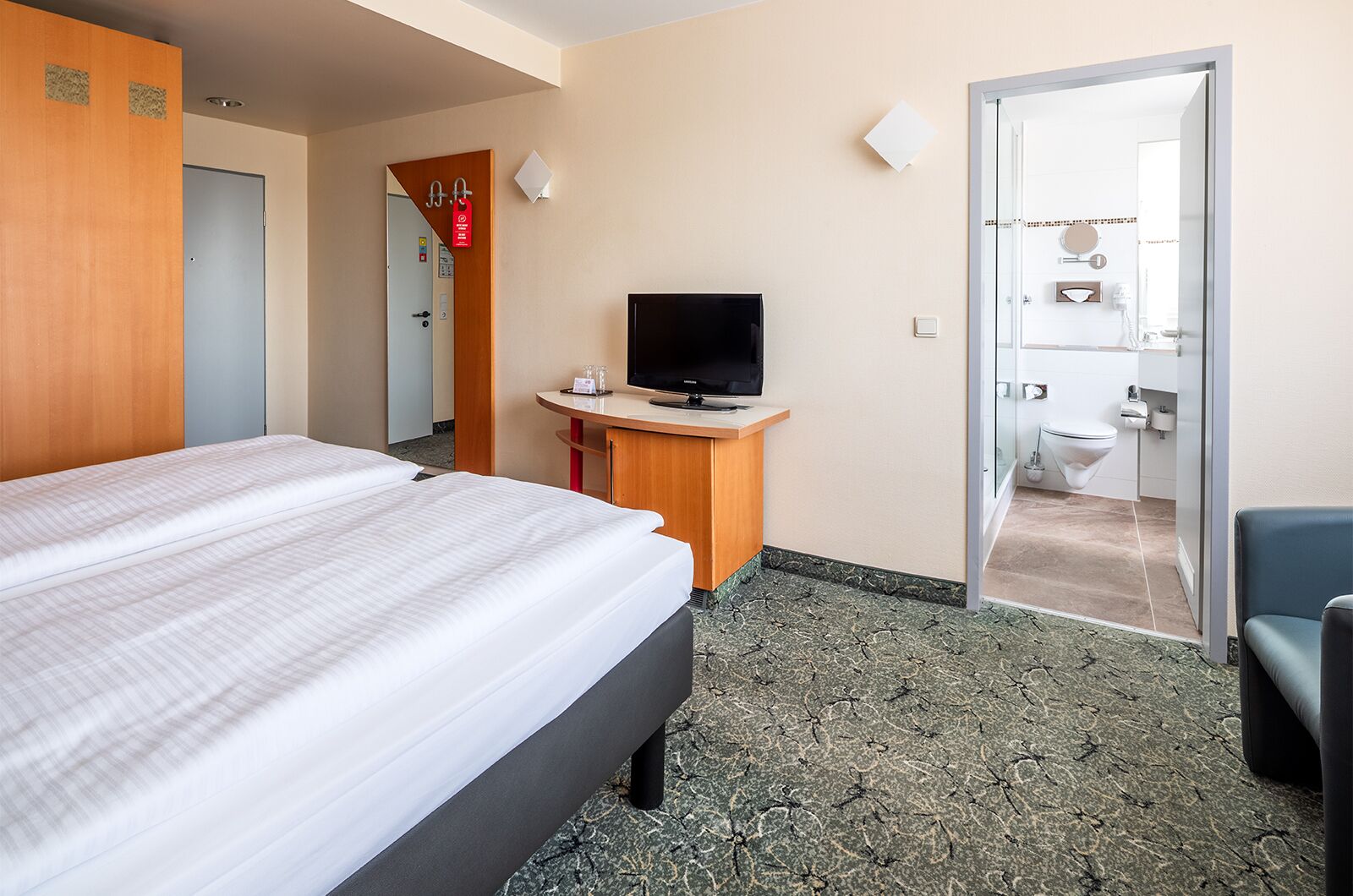 Ein Hotelzimmer mit Bett, Fernseher, Schreibtisch und einem durchgehenden Türspalte sichtbaren Badezimmer.