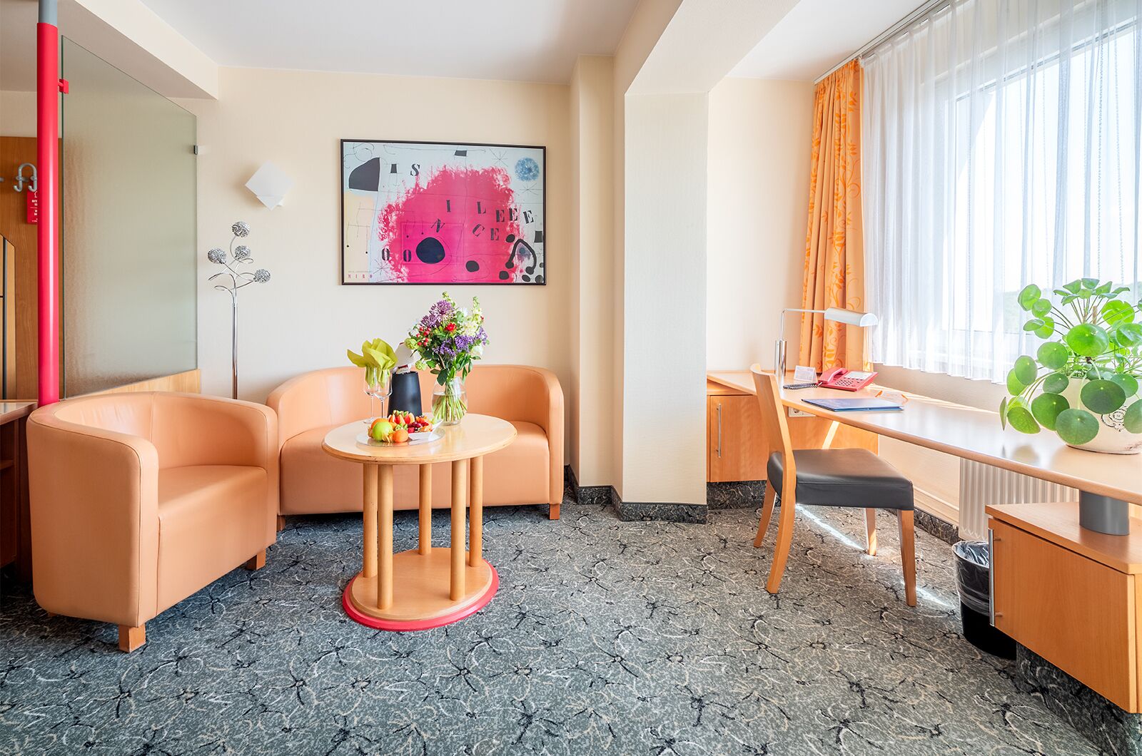 Quarto de hotel com sofá, uma pequena mesa redonda com um vaso de flores, escrivaninha com cadeira, uma janela grande com cortinas e um quadro na parede.