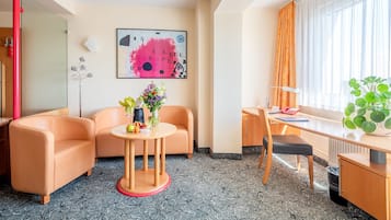Quarto de hotel com sofá, uma pequena mesa redonda com um vaso de flores, escrivaninha com cadeira, uma janela grande com cortinas e um quadro na parede.