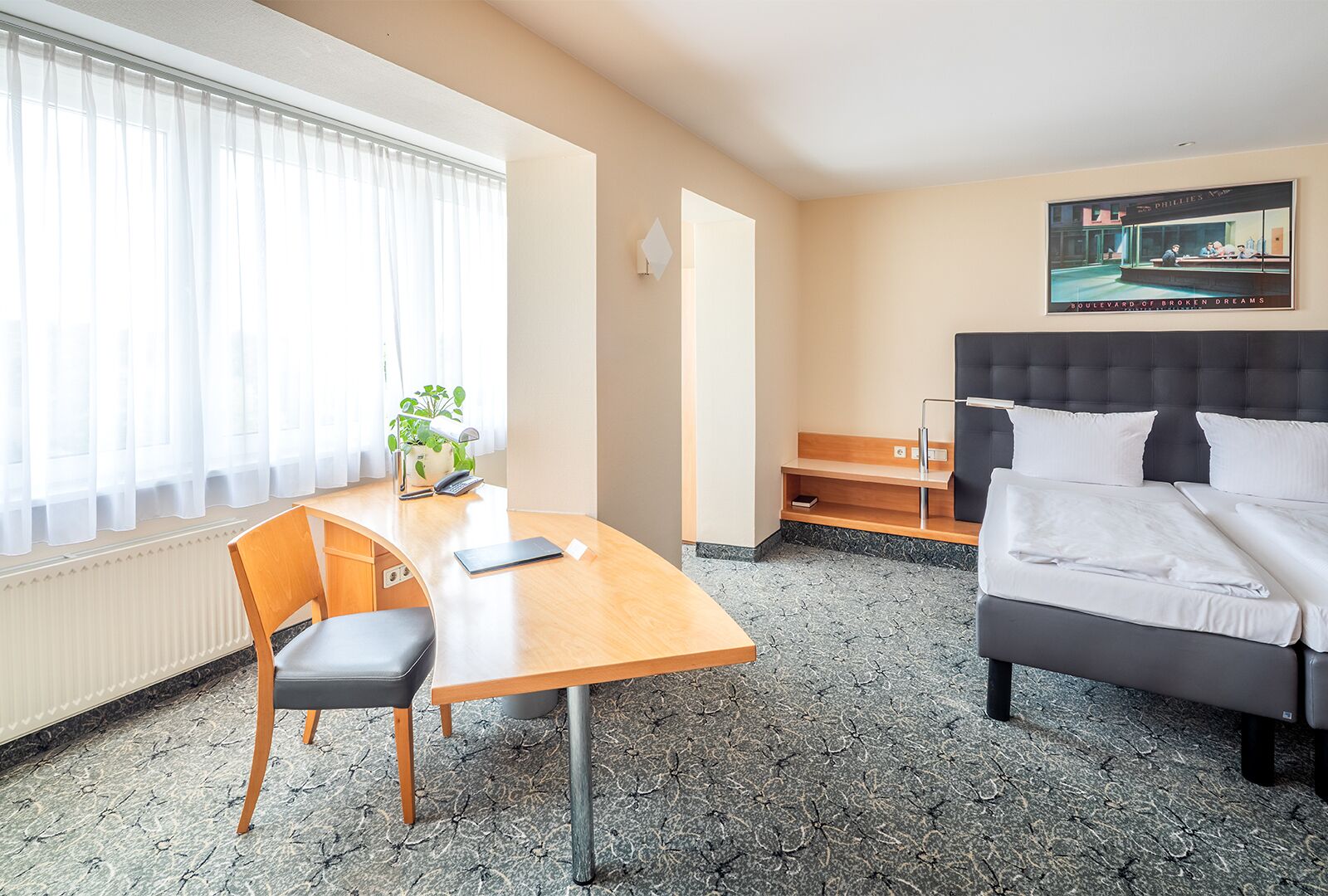 Habitación de hotel con cama, escritorio, una silla, una planta y un cuadro en la pared.