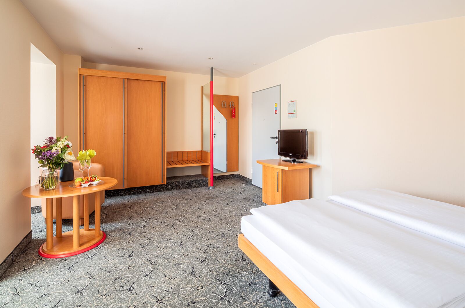 Ein Hotelzimmer mit einem Bett, einem Fernseher, einem Schreibtisch mit Blumenvase, einem Holzschrank und einer roten Stange.