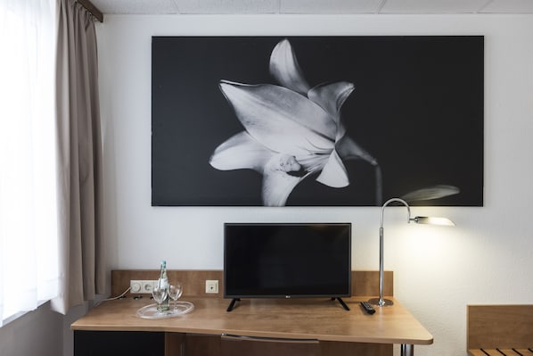 Habitación de hotel con escritorio de madera, televisor de pantalla plana, un cuadro de flor colgado en la pared y una lámpara de noche.