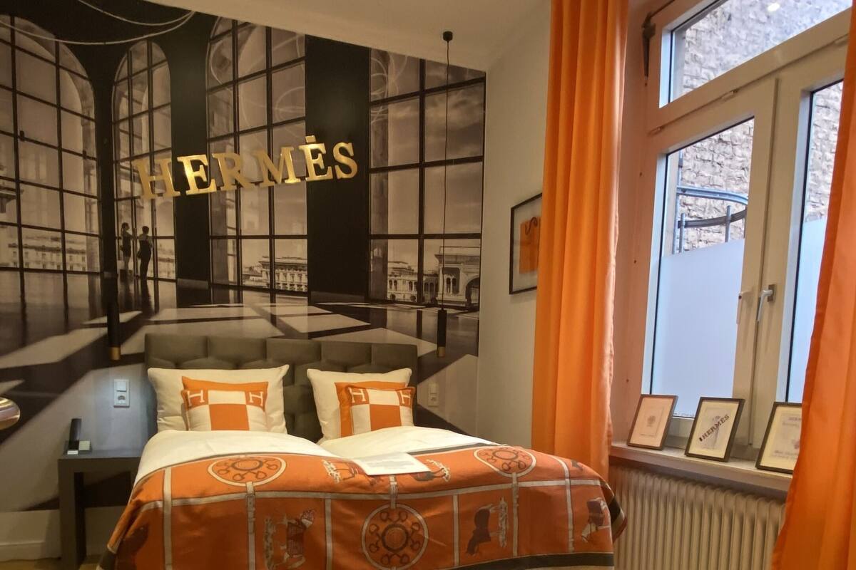 A bedroom with a large bed, a window with orange curtains, and a wall with 'HERMES' signage.